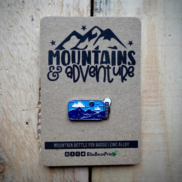 Mountain-themed bottle pin badge on a cardboard backing with 'Mountains & Adventure' text.