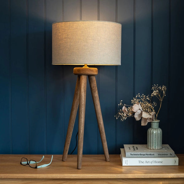 Wooden tripod lamp with beige shade on a wooden surface against a blue wall.