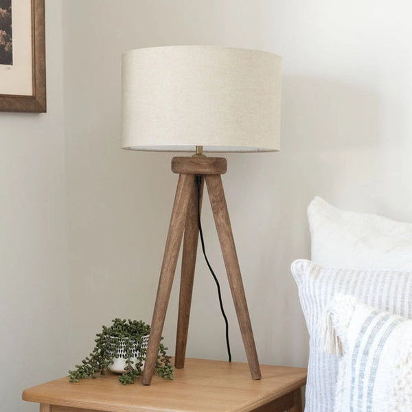 Wooden tripod lamp with a white shade on a wooden side table next to a bed.