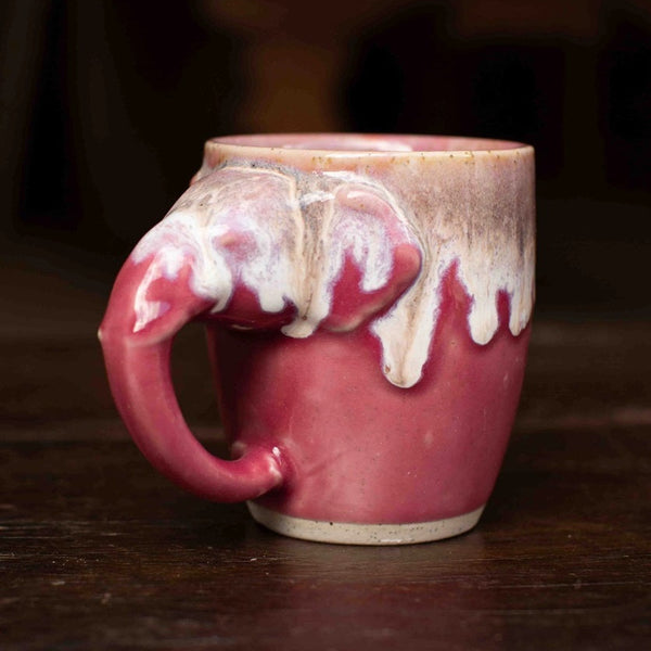 A handmade ceramic mug with a unique handle shaped like a curled piece of clay, featuring a dusty rose pink glaze with white dripping glaze accents on a dark wooden table.