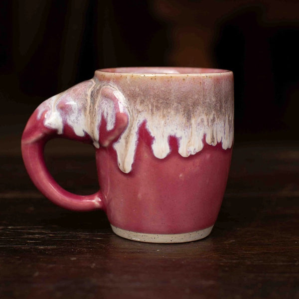 A handmade ceramic mug with a glossy pink and white dripping glaze, resting on a dark wooden surface.