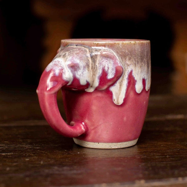 A handmade ceramic mug with a sculptural elephant handle, featuring a glossy pink body, white dripping glaze, and a rustic brown base, set on a dark wooden table.