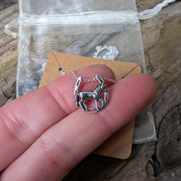 Silver horse necklace held between fingers with a wooden background