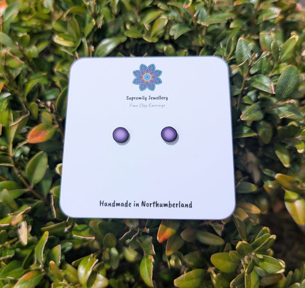 Two small oval stud earrings in a deep purple shade, mounted on a white display card from 'Supremely Jewellery', set against a background of green holly leaves.