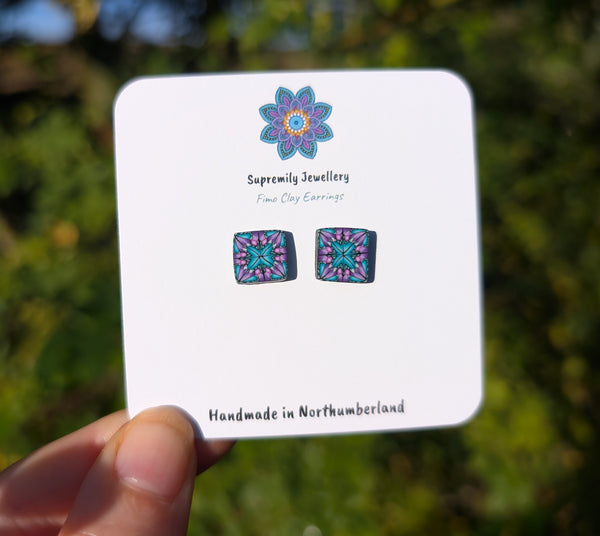 A hand holds a white display card featuring a pair of square stud earrings with a colorful mandala design. The card is branded 'Supremily Jewellery' and has a blurred green outdoor background.