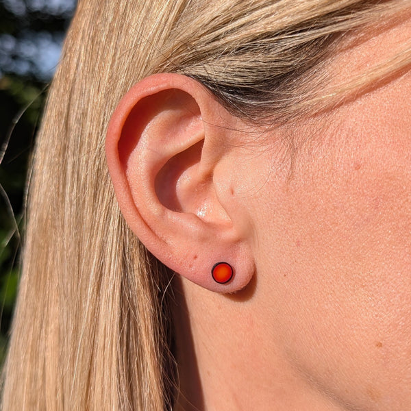 A close-up of a person's ear wearing a small round stud earring with a bright orange-red center and a darker red outer ring, set against blonde hair and a blurred outdoor background.