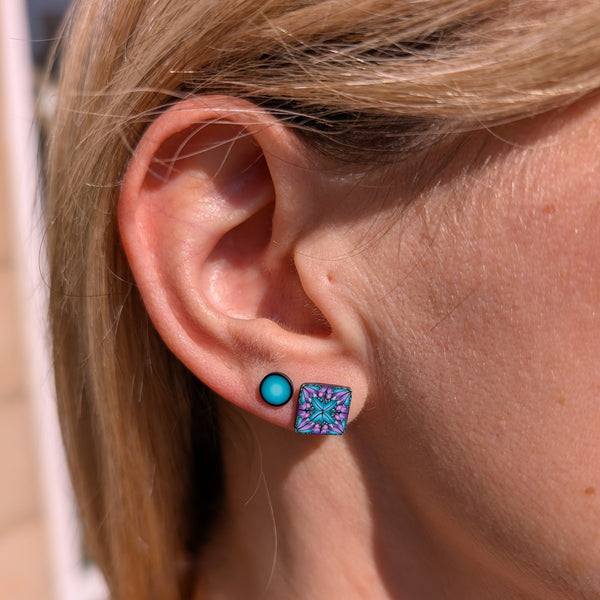 A close-up of a person's ear wearing two earrings: a small round turquoise stud and a square stud with a colorful mandala-like pattern in blue, purple, and pink.