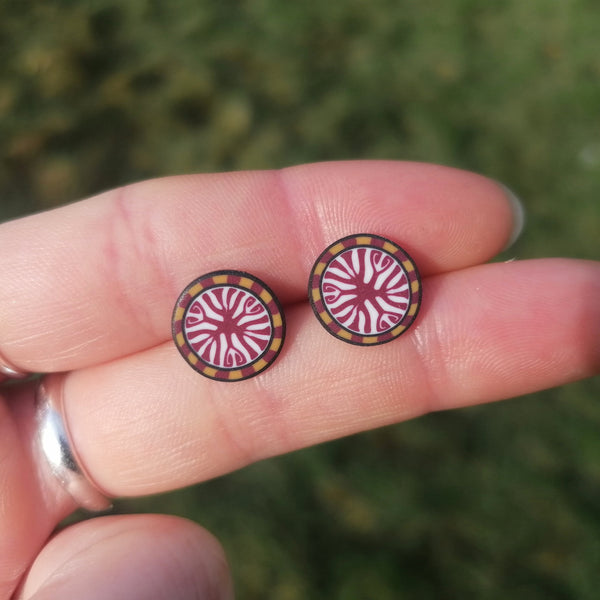 Round earrings with a red and gold design held between fingers against a blurred green background