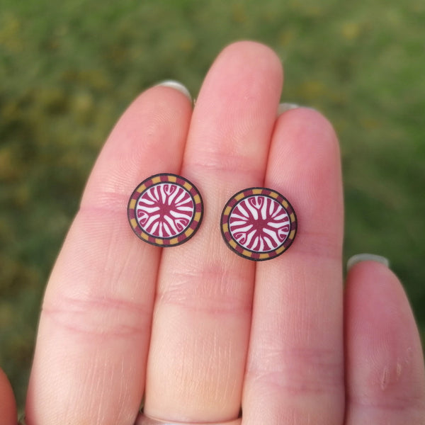 Pair of round earrings with a pink and black design held between fingers against a blurred green background