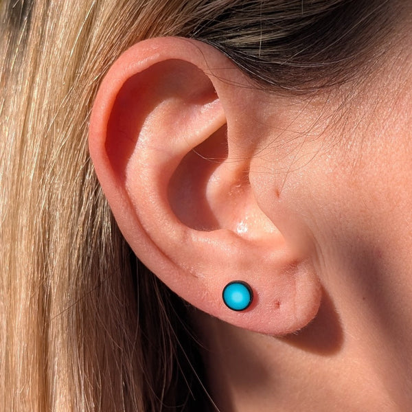 A close-up of a person's ear wearing a small round stud earring in bright turquoise blue, with blonde hair and skin visible in natural sunlight.