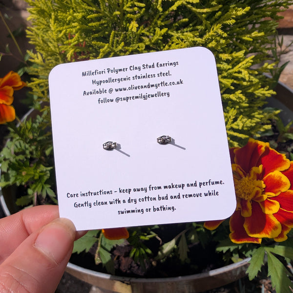 Text on a white card with the back of stud earrings showing. Held by fingers with a green leafy background and orange flowers.