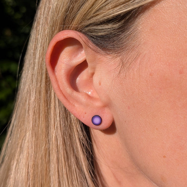 A close-up of a person's ear wearing a small round stud earring with a purple gradient design, set against natural outdoor background.