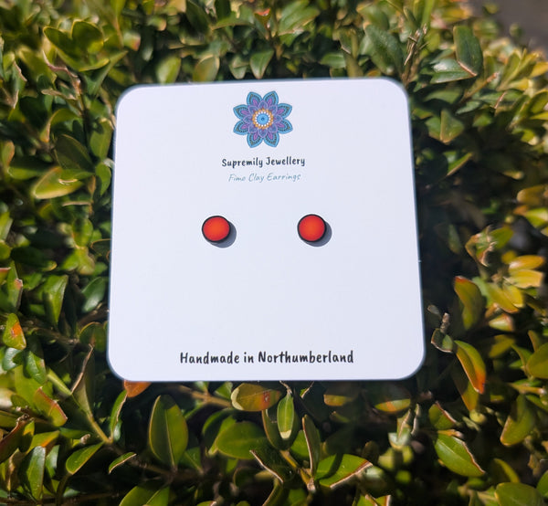 A pair of small oval stud earrings with red centers and black outer edges, displayed on a white card labeled 'Supremily Jewellery' against a background of green leaves.
