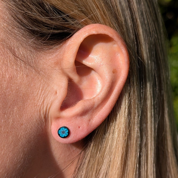 A close-up of a person's ear wearing a small round stud earring with a bright blue floral or starburst design on a dark background.
