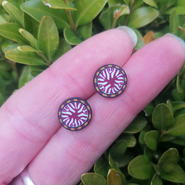 Round earrings with a colorful design held between fingers against a green leafy background
