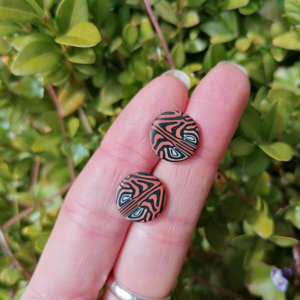Hand holding two decorative earrings with a leaf pattern against a green leafy background