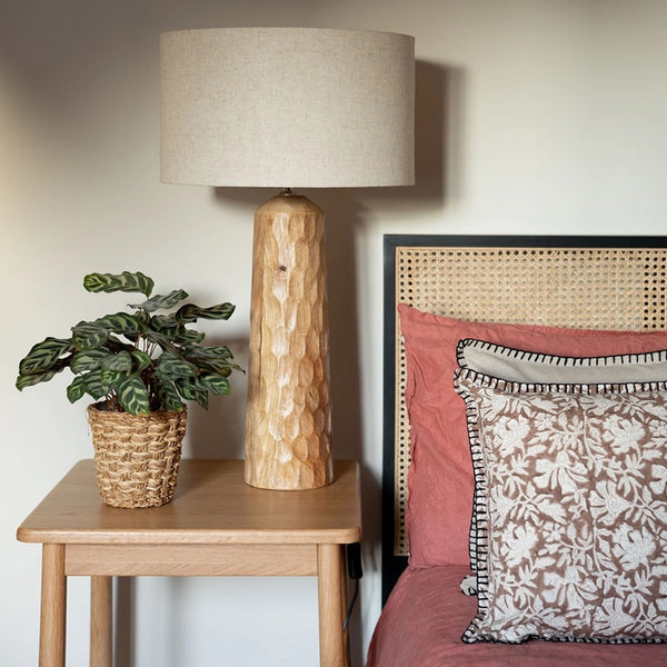 Wooden table with a lamp and plant next to a bed with floral and solid-colored pillows.