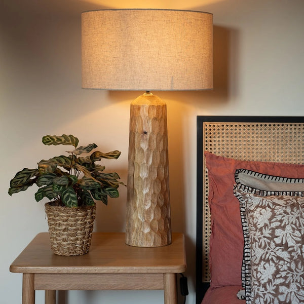 Wooden lamp on a side table next to a bed with pillows and a plant.