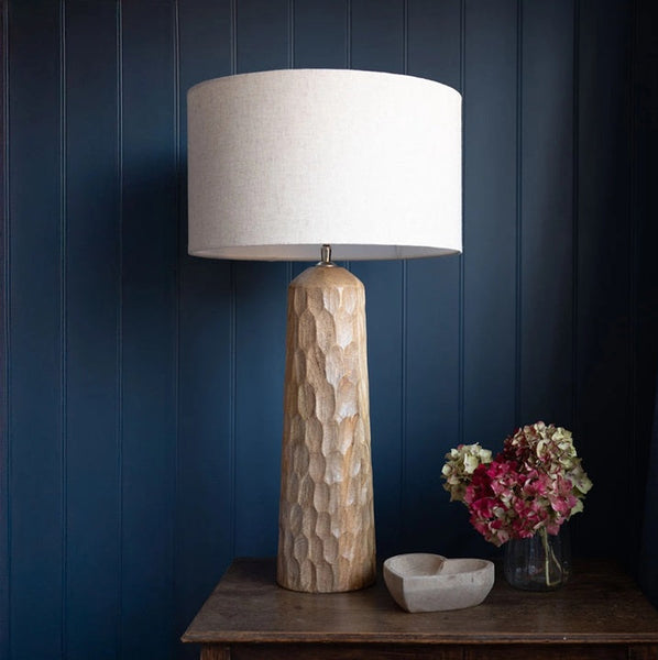 Decorative lamp with textured base and white shade on a wooden table against a dark blue wall.