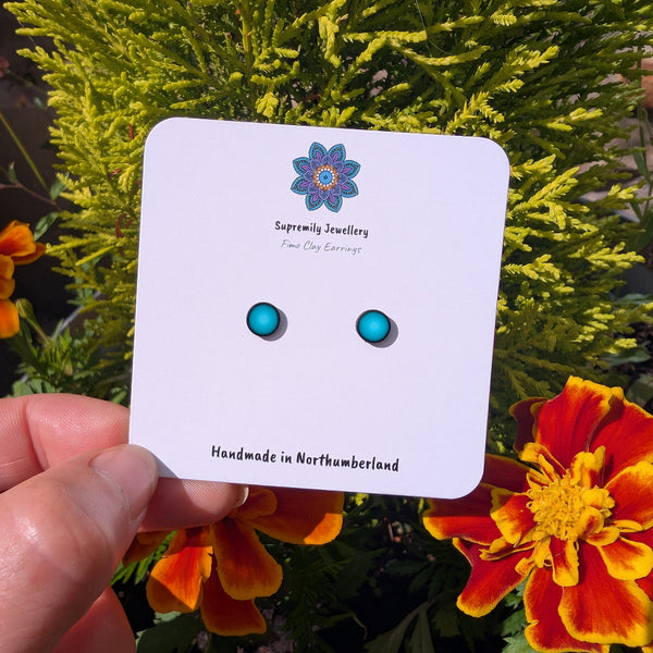 A small pair of vibrant teal green polymer clay stud earrings on a white card with Supremily Jewellery logo held in fingers against a green lefy background with ornage flowers.