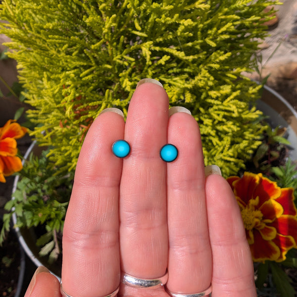 A small pair of vibrant teal green polymer clay stud earrings held between fingers with a green leafy flowery background.