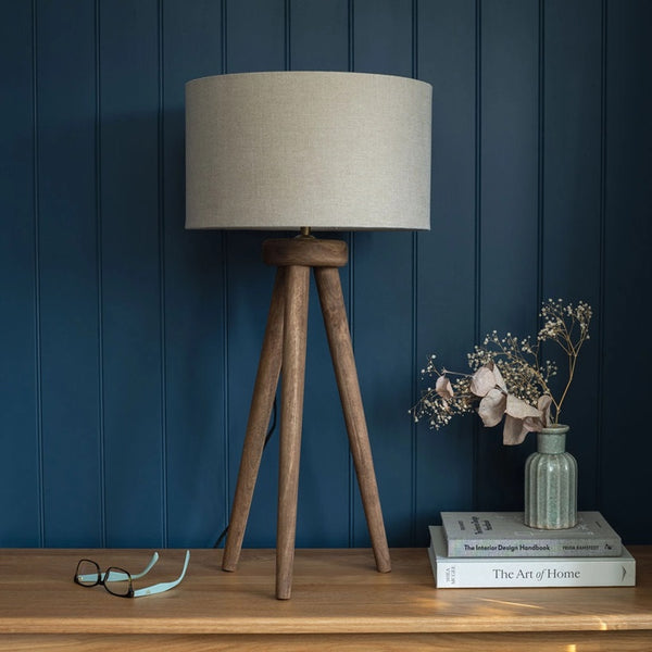 Table lamp with wooden base and beige shade on a wooden surface against a blue paneled wall.