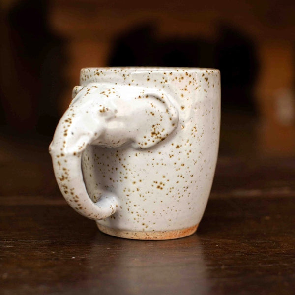 A handmade ceramic mug featuring a sculpted elephant handle, finished in a speckled off-white glaze with brown and gold flecks, resting on a dark wooden table.