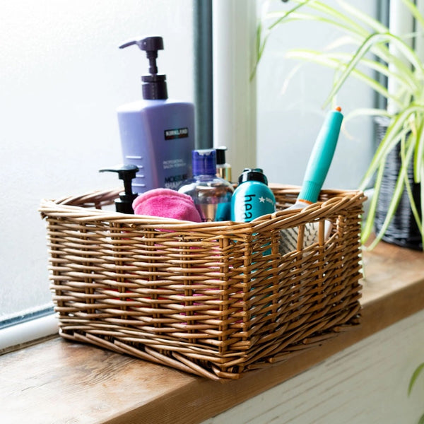 A natural wicker storage basket filled with toiletries, including a purple pump bottle, a pink sponge, and a turquoise toothbrush, sitting on a wooden windowsill.