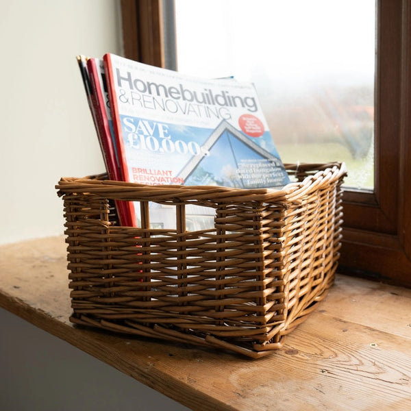 A rectangular woven wicker basket sits on a wooden windowsill next to a window, holding several magazines.