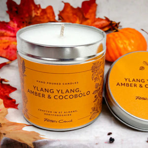 A Ylang Ylang, Amber & Cocobolo scented candle from Potters Crouch, displayed in a cylindrical orange tin with a lid, surrounded by autumn leaves.