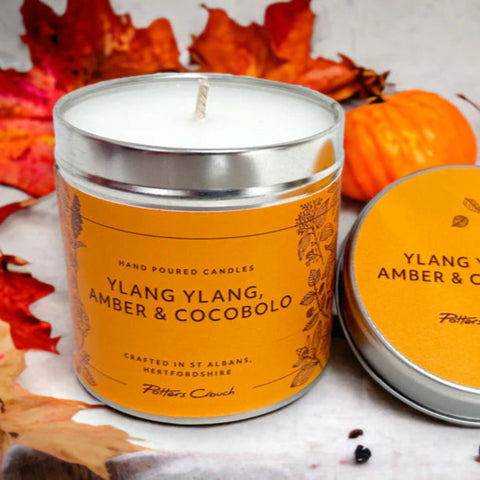A Ylang Ylang, Amber & Cocobolo scented candle from Potters Crouch, displayed in a cylindrical orange tin with a lid, surrounded by autumn leaves.